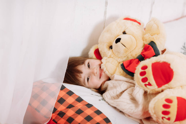 Adorable smiling little girl is lying and playing with teddy bear on bed in room decorated garlands for New Year. Winter weekends. Cozy scene. Christmas holiday atmosphere - Photo, Image