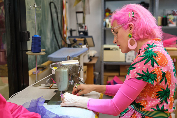 Rózsaszín hajjal és színes ruhával rendelkező szabó menetvágó gép varróműhelyben. - Fotó, kép