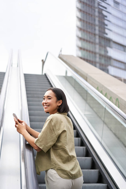Young asian girl going up on an escalator, holding smartphone, smiling while walking in city. Copy space - Foto, Imagem