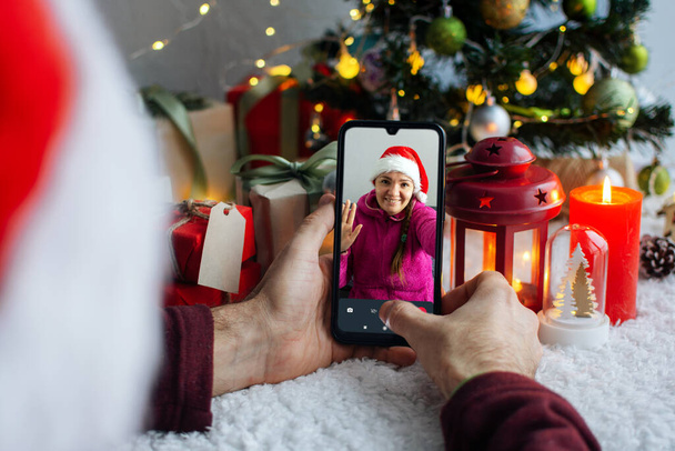 クリスマス・イブのビデオ通話だ。男は親戚や友人をスマートフォンで呼び出す。少女はビデオ通話に答えて手を振った。背景にはクリスマスツリーと装飾があります。. - 写真・画像