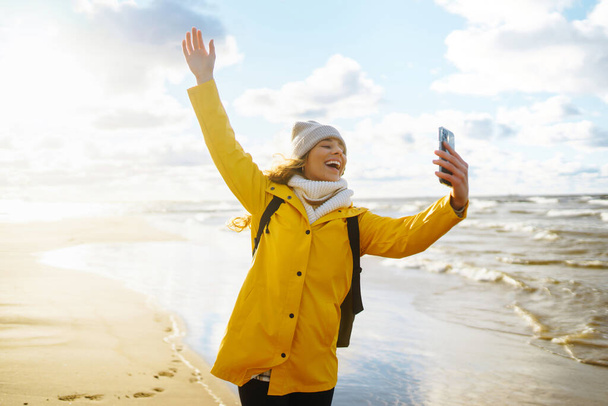 Young tourist with phone. Selfie time. Tourist in a yellow jacket posing by the sea at sunset. Travelling, lifestyle, adventure. - Foto, Imagem