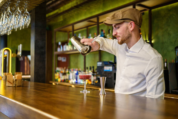 Człowiek z brodą w białej koszuli i czapce wypełniającej szklankę koktajlem z shakera stojąc za ladą i pracując w barze - Zdjęcie, obraz