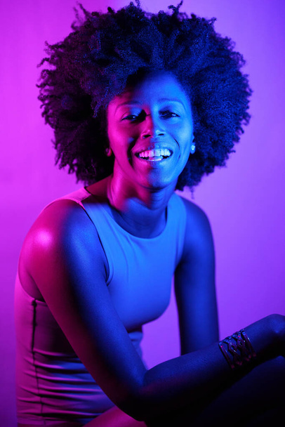 Heureuse femme afro-américaine aux cheveux bouclés souriant et regardant la caméra sous la lumière violette au néon - Photo, image