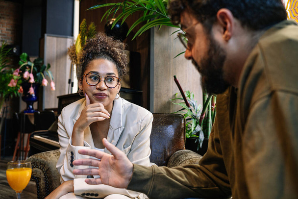 Black woman in smart casual clothes touching chin and listening to bearded coworker while sitting in armchair during business meeting in restaurant - 写真・画像