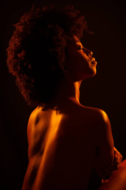 Afro saç stili kapalı gözleri olan ve siyah arka planda parlak neon turuncu ışığın tadını çıkaran çıplak Afro-Amerikan kadın modeli. - Fotoğraf, Görsel