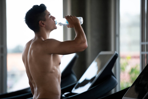 Молодой кавказский здоровый спортсмен пьет воду в спортзале после тренировки на беговой дорожке. - Фото, изображение