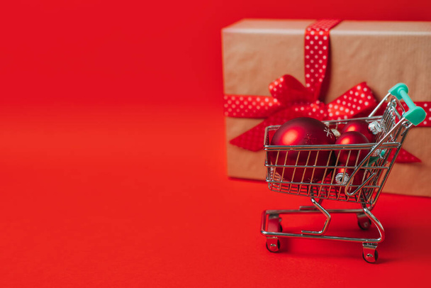 クリスマスセール赤い背景にギフトボックスと赤いクリスマスの装飾ボールとミニトロリーカートとショッピングウェブバナー。ミニマルクリスマスショッピング販売ウェブバナーコピースペースコンセプト - 写真・画像