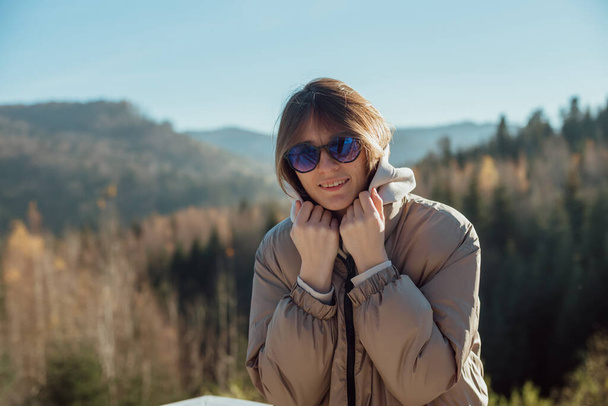 Een meisje in warme kleren staat op het terras tegen de achtergrond van de bergen; meisje in een zonnebril; houdt de kap met zijn handen - Foto, afbeelding