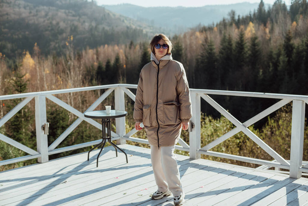 девушка в теплой одежде стоит на террасе на фоне гор; девушка в солнечных очках; гуляет к камере на белой террасе - Фото, изображение