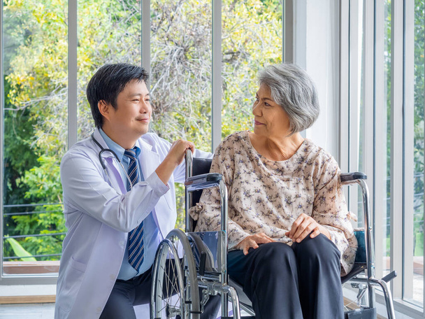 Улыбающийся позитивный азиат врач в белом халате говорит с заботой о пожилой пациентке старшего возраста, которая сидит в инвалидной коляске в медицинском кабинете в больнице. Кардиология, здравоохранение и медицинская концепция. - Фото, изображение