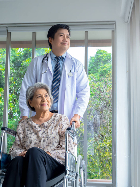 Gülümseyen mutlu Asyalı yaşlı yaşlı kadın hasta hastanede dikey bir şekilde beyaz önlüklü erkek doktorla birlikte tekerlekli sandalyede oturuyor. Bakım, sağlık ve tıbbi konsept. - Fotoğraf, Görsel