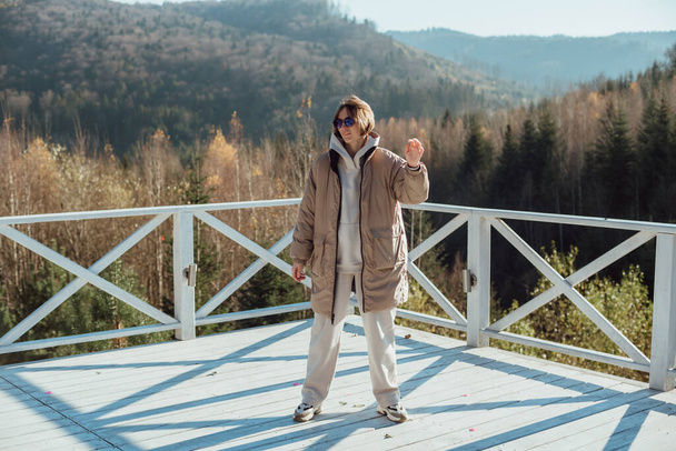 una chica en ropa de abrigo se para en la terraza sobre el fondo de las montañas; chica en gafas de sol; la mano izquierda se levanta - Foto, Imagen