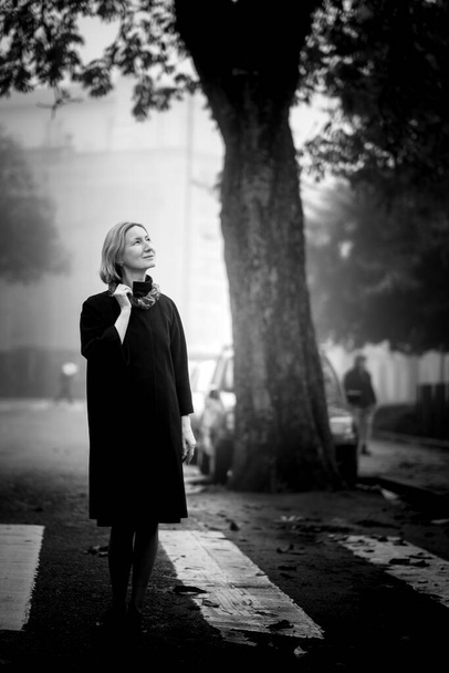 Una mujer con un abrigo de otoño está en la acera de la ciudad. Foto en blanco y negro. - Foto, Imagen