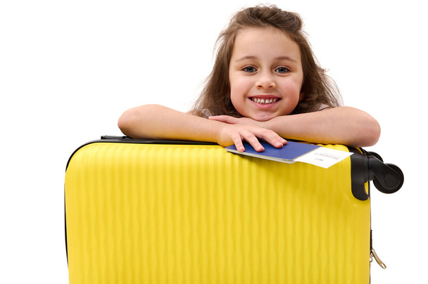 Szczęśliwa mała pasażerka z kartą pokładową, słodko uśmiechnięta patrząca w kamerę, opierająca się na żółtej walizce, odizolowana na białym tle. Podróż. Podróż. Podróż. Dzieci podróżują. Powierzchnia reklamowa - Zdjęcie, obraz