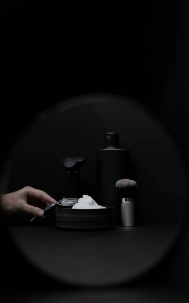 Shaving foam in a bowl, a shaving brush, an electric razor, a bottle of aftershave cream and a female hand holding a razor in the reflection of a round mirror on a black background, close-up side view. The concept of a male hairdresser, beauty salon, - Photo, Image
