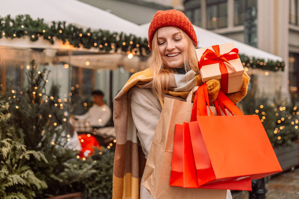 Boldog fiatal vonzó nő papírzacskókkal és ajándékdobozokkal a kezében karácsonyi bevásárlás után a plázában. Vidám gyönyörű lány tartsa bevásárló táskák és jelen dobozok a kezében. Téli ünnepek értékesítése - Fotó, kép