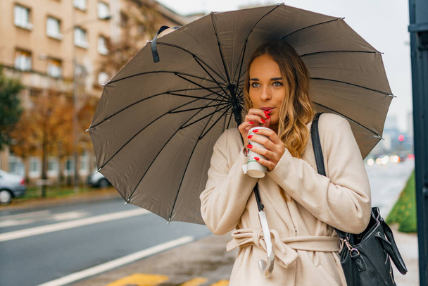 Облачный день, дождливая погода осенью женщины держат ее горячий кофе чашку, как она ждет общественного транспорта и пьет кофе - Фото, изображение