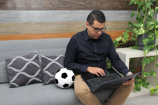 Λατίνος ενήλικας υπάλληλος γραφείου παρακολουθεί ποδοσφαιρικούς αγώνες στο laptop εργασίας του κατά τη διάρκεια των ωρών γραφείου το πρωί, τον βλέπει νευρικό, λυπημένος, αγχωμένος, θυμωμένος δίπλα στην μπάλα ποδοσφαίρου του - Φωτογραφία, εικόνα