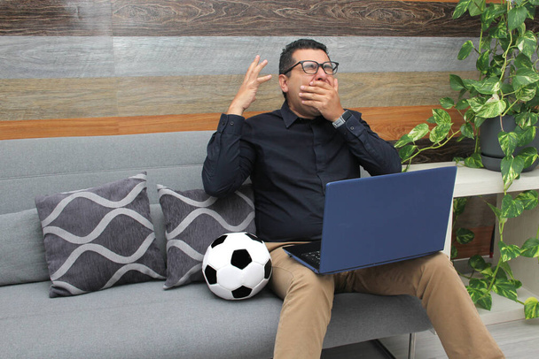 Латиноамериканський службовець спостерігає за футбольними іграми на своєму робочому ноутбуку в робочий час вранці, він бачить його нервовим, сумним, стресовим, сердитим поруч з футбольним м'ячем. - Фото, зображення