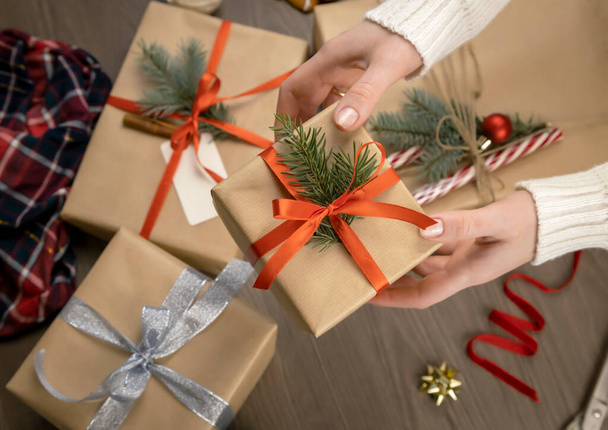Noel süslemesinin arka planında kırmızı kurdeleli ve Noel ağacı dallı hediye kutusu olan kadın eller. Tatil için hediye hazırlama konsepti. - Fotoğraf, Görsel