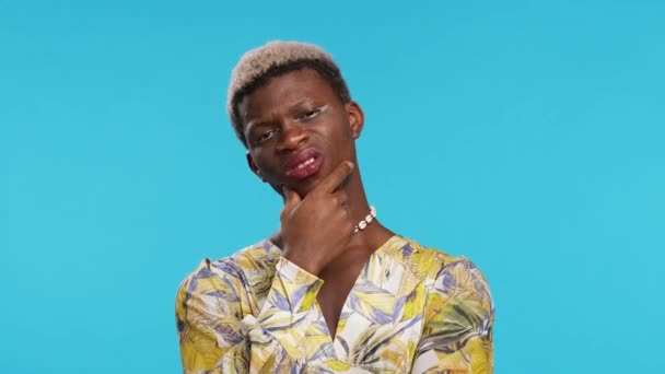 Скептик черный трансгендер в стильном платье с рукой на поясе касаясь подбородка и глядя вверх, размышляя над проблемой на синем фоне - Кадры, видео
