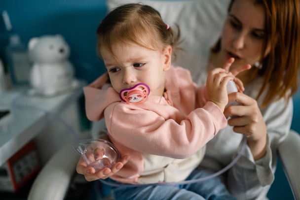One toddler girl small caucasian daughter sitting in chair with her mother refuse to use nebulizer at home child using vapor steam inhaler mask inhalation medical procedure medicament treatment - Photo, Image