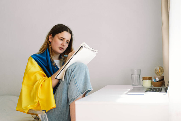 Uma adolescente ucraniana está envolvida na bandeira ucraniana e está desenhando ou fazendo trabalhos de casa no quarto. Um adolescente estuda em casa. O conceito de estudos durante a guerra na Ucrânia. - Foto, Imagem