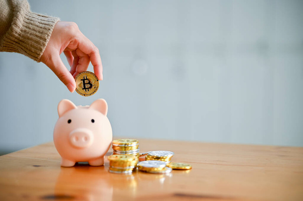 Eine weibliche Hand hält eine goldene Bitcoin, steckt Münzen in ein Sparschwein, stapelt Goldmünze und Sparschwein auf einem Holztisch. Investition, Sparen, Finanzkonzept - Foto, Bild