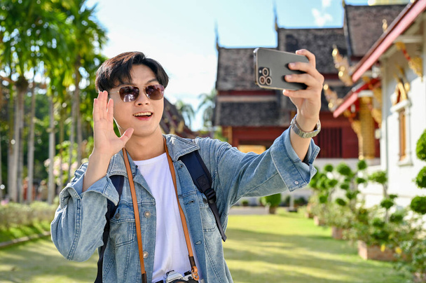 Guapo y alegre joven asiático viajero masculino con gafas de sol, tomar selfie o hacer un video vlog con su teléfono inteligente en el hermoso templo de Tailandia. - Foto, imagen