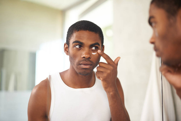 Hij zorgt voor zijn huid. Een jonge man die crème op zijn gezicht aanbrengt terwijl hij in de spiegel kijkt - Foto, afbeelding