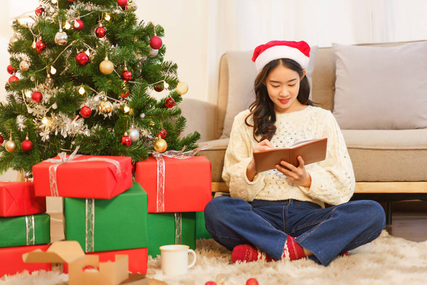 Χριστούγεννα έννοια εορτασμού, Ασιάτισσα γυναίκα κάθεται στο πάτωμα κοντά στο χριστουγεννιάτικο δέντρο για να γράψει στο σημειωματάριο. - Φωτογραφία, εικόνα