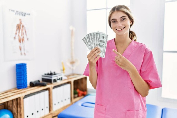 Молодая женщина-физиотерапевт, работающая в клинике болеутоляющих, держит в руках доллары, улыбаясь, показывая пальцем и рукой.  - Фото, изображение