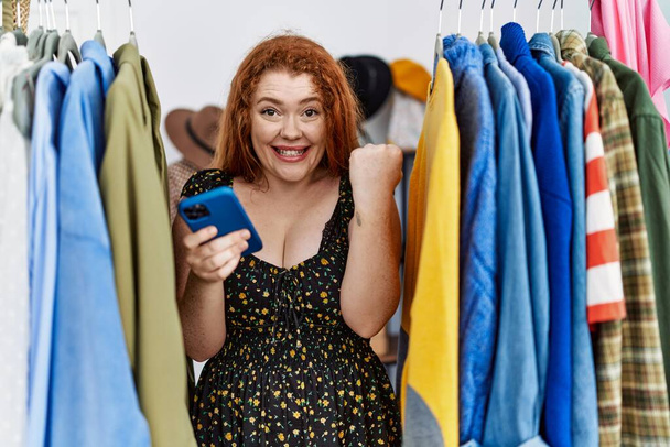 Young redhead woman searching clothes on clothing rack using smartphone screaming proud, celebrating victory and success very excited with raised arms  - Фото, зображення