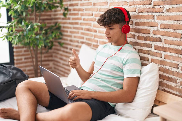 Ισπανόφωνος έφηβος χρησιμοποιώντας φορητό υπολογιστή στο σπίτι κάθεται στην κρεβατοκάμαρα χαμογελώντας χαρούμενος και θετικός, αντίχειρας επάνω κάνει εξαιρετική και την έγκριση σημάδι  - Φωτογραφία, εικόνα