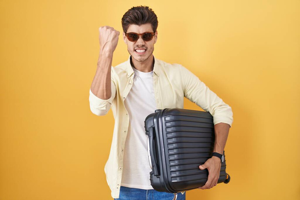 Joven hombre hispano sosteniendo maleta que va de vacaciones de verano enojado y loco levantando el puño frustrado y furioso mientras grita con ira. rabia y concepto agresivo.  - Foto, Imagen