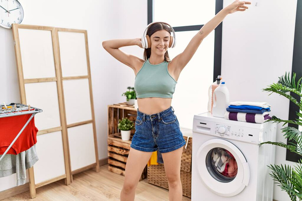 Young hispanic woman listening to music waiting for washing machine at laundry room - Photo, Image