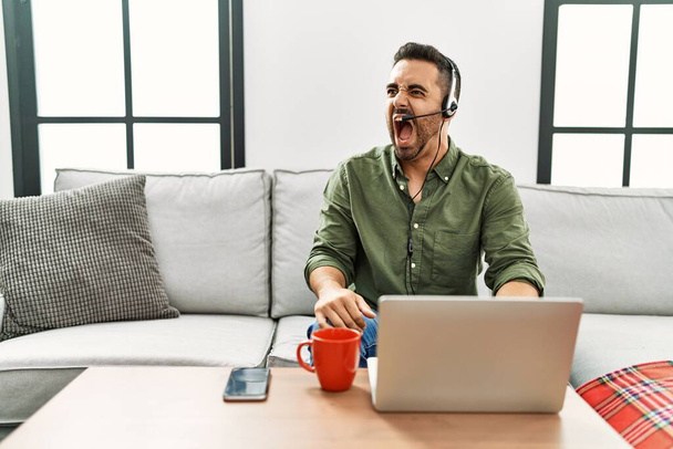 Jonge Spaanse man met baard die een callcenter agent headset draagt die thuis werkt boos en gek schreeuwend gefrustreerd en woedend, schreeuwend van woede. woede en agressief concept.  - Foto, afbeelding