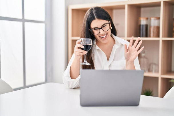 jonge brunette vrouw doen videogesprek met laptop drinken van een glas wijn kijken positief en gelukkig staan en glimlachen met een zelfverzekerde glimlach tonen tanden  - Foto, afbeelding