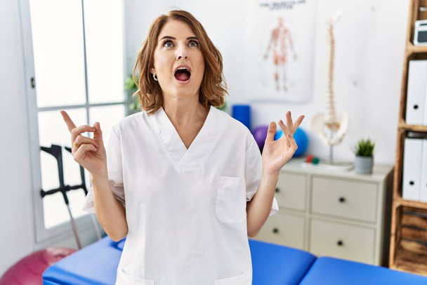 Orta yaşlı fizyoterapist kadın ağrı kesici kliniğinde çalışıyor. Şaşırmış ve şaşırmış bir şekilde yukarı bakıyor ve parmaklarıyla işaret ediyor ve kollarını kaldırıyor..  - Fotoğraf, Görsel