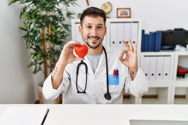 Νεαρός γιατρός κρατώντας την καρδιά στην ιατρική κλινική κάνει ok υπογράψει με τα δάχτυλα, χαμογελώντας φιλικό gesturing εξαιρετικό σύμβολο  - Φωτογραφία, εικόνα