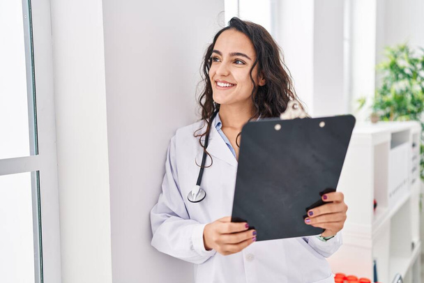 Young hispanic woman wearing doctor uniform holding clipboard at clinic - Photo, image