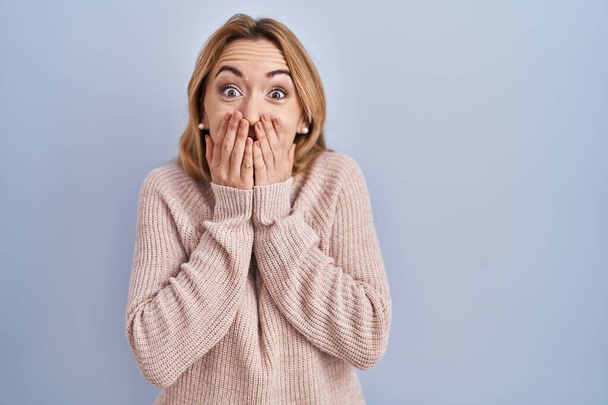 Spaanse vrouw staat over blauwe achtergrond lachen en beschaamd giechelen bedekken mond met handen, roddel en schandaal concept  - Foto, afbeelding