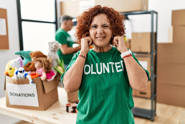 Frau mittleren Alters, die am Spendenstand ein freiwilliges T-Shirt trägt und die Ohren mit Fingern bedeckt, die sich über den Lärm lauter Musik ärgern. Gehörloses Konzept.  - Foto, Bild