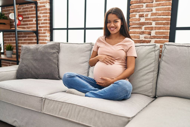 Young latin woman pregnant touching belly sitting on sofa at home - Photo, Image