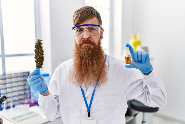 Vörös hajú férfi hosszú szakállal, a tudós laboratóriumban dolgozik, füvet tart és cbd olaj szkeptikus és ideges, rosszalló feldúlt a probléma miatt. negatív személy.  - Fotó, kép