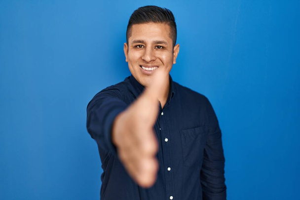 Der hispanische junge Mann steht vor blauem Hintergrund und lächelt freundlich und reicht zur Begrüßung Handschlag. Erfolgreiches Geschäft.  - Foto, Bild
