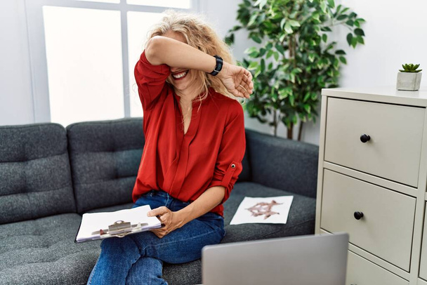Женщина-психолог средних лет в консультационном кабинете, улыбаясь, весело играя, подглядывает в буу с руками, показывающими лицо. удивленные и ушедшие  - Фото, изображение