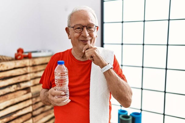 Starszy mężczyzna ubrany w odzież sportową i ręcznik na siłowni, wyglądający pewnie przed kamerą, uśmiechnięty ze skrzyżowanymi rękami i podniesioną ręką na brodzie. pozytywne myślenie.  - Zdjęcie, obraz