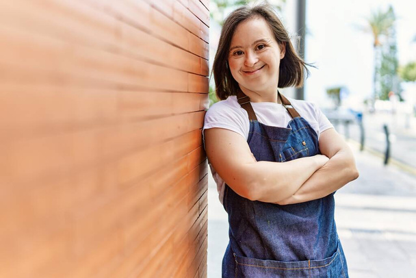 Young down syndrome woman smiling confident wearing apron at street - 写真・画像