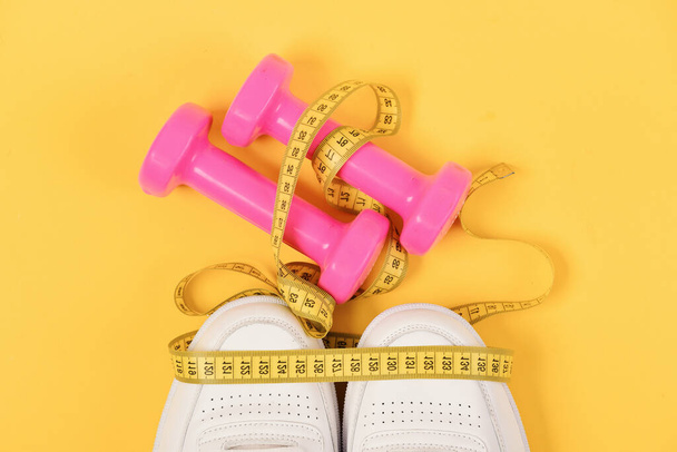Sports and healthy lifestyle concept. Centimeter in yellow color on trainers and pink dumbbells, top view. Sneakers with measuring tape on yellow background. Sport shoes and equipment for fit shape - Foto, afbeelding